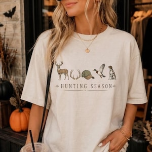 May include: A cream-colored short-sleeve t-shirt featuring a graphic print of a deer, antlers, a green baseball cap, a flying duck, and a dog. The text "HUNTING SEASON" is printed in black lettering below the illustrations with decorative arrows.