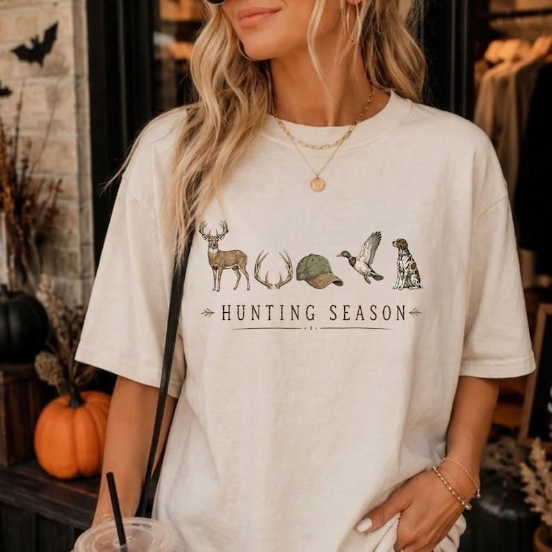 May include: A cream-colored short-sleeve t-shirt featuring a graphic print of a deer, antlers, a green baseball cap, a flying duck, and a dog. The text "HUNTING SEASON" is printed in black lettering below the illustrations with decorative arrows.