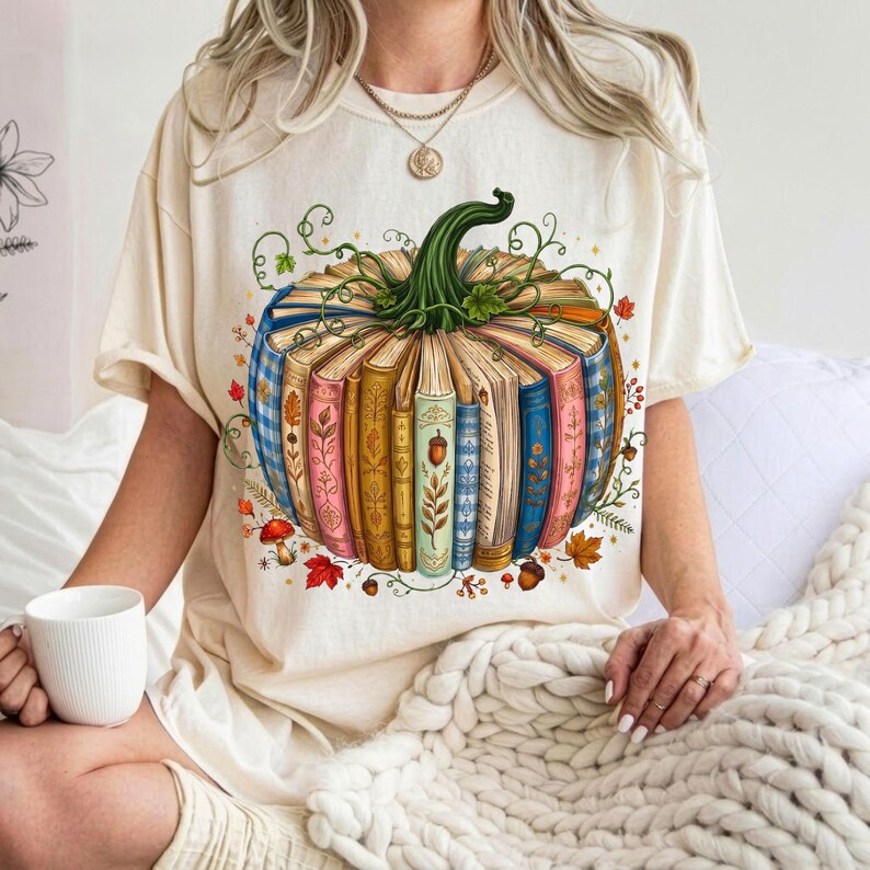 May include: A cream-colored t-shirt featuring a graphic of a pumpkin constructed from a circular arrangement of colorful books. The pumpkin has a green stem and is surrounded by small illustrations of autumn leaves and mushrooms.