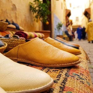 Puede incluir: Un par de babuchas de cuero color amarillo mostaza con punta afilada y suela plana, exhibidas junto a otro calzado de cuero en varios colores, incluyendo crema y azul oscuro, con costuras visibles a lo largo de los bordes de las suelas.