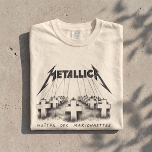Może przedstawiać: Kremowy t-shirt z krótkim rękawem z czarną grafiką przedstawiającą logo Metallica z nitkami przyczepionymi do pola krzyży. Poniżej grafiki znajduje się czarny napis "MAÎTRE DES MARIONNETTES". Koszulka posiada metkę marki Comfort Colors.