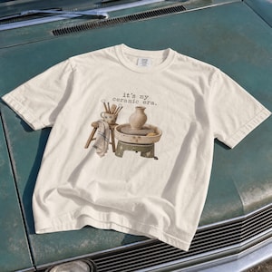 Op de afbeelding: Een off-white t-shirt met korte mouwen en een grafische print van een pottenbakkersschijf, een keramische vaas en gereedschap. De tekst "it's my ceramic era." staat in een eenvoudig zwart lettertype boven de illustratie gedrukt.