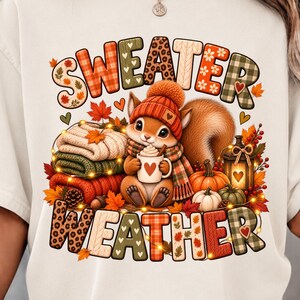 May include: A cream-colored t-shirt featuring a graphic of a squirrel wearing an orange beanie and scarf, holding a mug with a heart. The text reads "SWEATER WEATHER" in patterned letters, surrounded by pumpkins, autumn leaves, and a lantern.