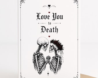 May include: A white greeting card featuring a black and white illustration of two skeletons holding hands. The text reads Love You to Death in a gothic font, accented by small red hearts and a decorative black border frame on a tan envelope.