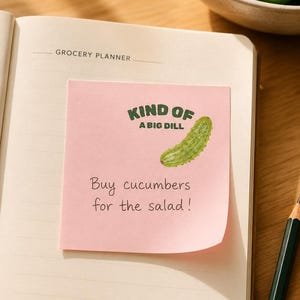 May include: A square pink sticky note featuring a green cucumber illustration and the text "KIND OF A BIG DILL" above the handwritten note "Buy cucumbers for the salad!" placed on the open page of a grocery planner notebook.