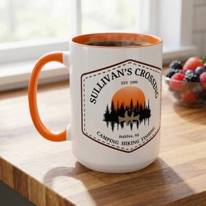Peut inclure: Un mug en céramique blanche avec un intérieur et une anse orange. L'avant présente un motif avec un coucher de soleil, des pins et un canoë. Le texte indique : Sullivan's Crossing, Est 1906, Halifax, NS, Camping, Hiking, Fishing, dans un hexagone.