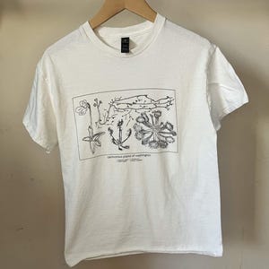 May include: A white short-sleeve t-shirt featuring a black line-art graphic of various plants. The print is enclosed in a rectangular border with the text "carnivorous plants of washington" and a numbered list of plant names printed below it.