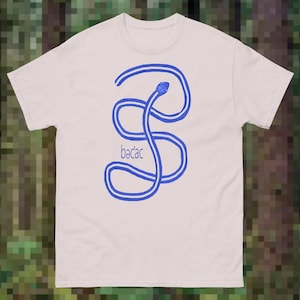 May include: A light-coloured short-sleeve t-shirt featuring a blue graphic of a coiled snake forming an S-shape. The text "bə'cəc" is printed in blue letters to the left of the snake graphic. The shirt has a crew neckline and a straight hem.