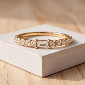 May include: A gold band ring featuring a central baguette-cut diamond flanked by a series of round brilliant-cut diamonds set along the top half of the band. The metal has a polished finish, and the stones are secured in a prong setting.