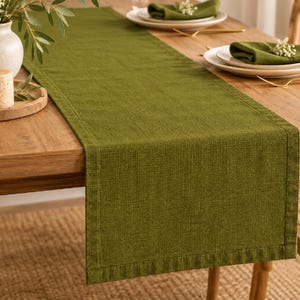 May include: A rectangular olive green table runner with a visible woven texture and stitched hemmed edges, draped lengthwise across a wooden dining table. The runner features a solid color and a structured fabric appearance.