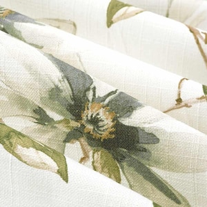 May include: Close-up of a floral fabric with a watercolor effect. The design features large, stylized flowers in shades of gray, green, and brown against a cream-colored background. The fabric is folded, showing the texture and pattern.