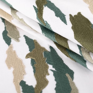Puede incluir: Una tela blanca con un patr&oacute;n de camuflaje bordado en tonos verde oscuro, verde oliva y beige claro. El bordado tiene una apariencia texturizada y elevada con formas irregulares y dentadas dispersas sobre el material base blanco.