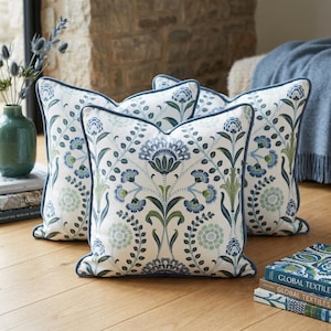 May include: A set of square throw pillows featuring a white fabric base with a repeating botanical pattern in shades of blue and green. Each pillow is finished with a solid blue piped edge, creating a cohesive look across the collection.