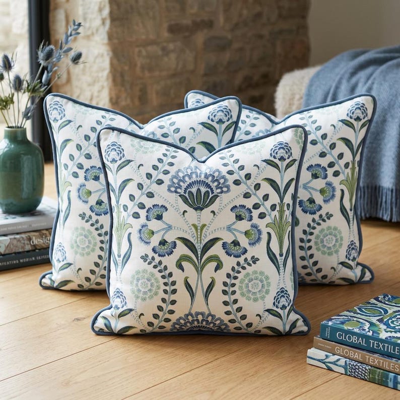 May include: A set of square throw pillows featuring a white fabric base with a repeating botanical pattern in shades of blue and green. Each pillow is finished with a solid blue piped edge, creating a cohesive look across the collection.