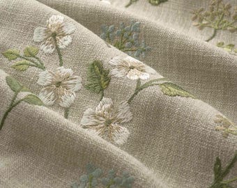 May include: A beige fabric featuring an embroidered floral pattern with white petals and green leaves. The textile has a visible woven texture and is draped in soft folds, showcasing the detailed stitching of the botanical design throughout.
