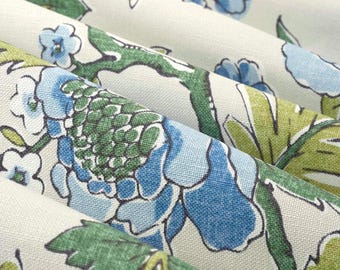 May include: A folded textile featuring a botanical print on a light beige background. The pattern consists of large blue flowers with dark green centers, smaller light blue blossoms, and vibrant green leaves with dark outlines on a woven fabric.