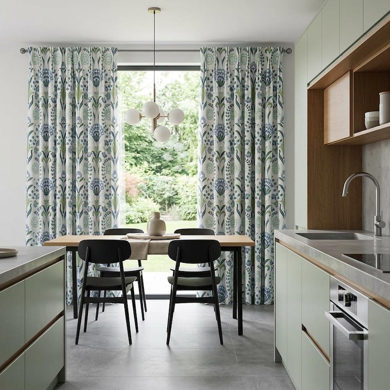 May include: Floor-length curtains featuring a repeating botanical pattern in shades of blue and green on a white background. The curtains hang from a metal rod, framing a large window that looks out onto a green garden area in a kitchen space.