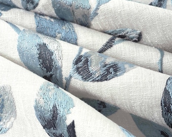 May include: A white fabric featuring an embroidered pattern of abstract leaf shapes in varying shades of blue. The embroidery uses a dense, textured stitch to create depth, while the base material has a visible woven texture throughout the surface.