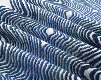 May include: A textile fabric featuring a repeating pattern of deep blue wavy lines embroidered onto a crisp white background. The fabric is gathered into soft, vertical folds, highlighting the textured, raised surface of the blue stitching.