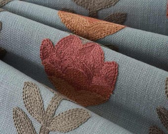May include: A light blue fabric featuring an embroidered floral pattern. The design includes rust-colored flower blossoms and brown leafy stems stitched directly onto the textured material, creating a repeating botanical motif across the surface.