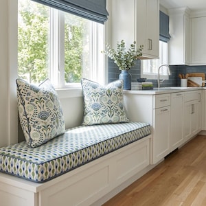May include: A window seat cushion and two square throw pillows featuring a blue, green, and white floral botanical pattern. The cushion and pillows are finished with blue piping and are placed on a white built-in bench in a kitchen.