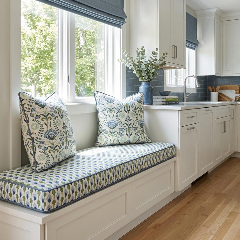 May include: A window seat cushion and two square throw pillows featuring a blue, green, and white floral botanical pattern. The cushion and pillows are finished with blue piping and are placed on a white built-in bench in a kitchen.