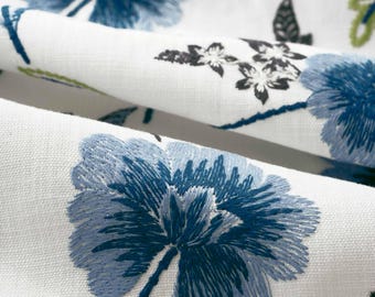 May include: A white fabric featuring detailed embroidery of large blue flowers with dark blue centers, smaller black floral motifs, and thin green stems. The textile has a visible woven texture and is folded to show the layered floral pattern.