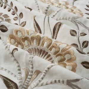 May include: Close-up of a white fabric with embroidered floral designs in brown and beige. The fabric has a subtle texture and a delicate, intricate pattern.
