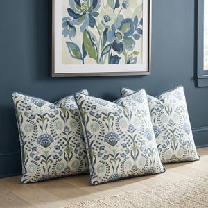 May include: Three square throw pillows featuring a white base with a repeating botanical pattern in shades of blue and sage green. Each pillow has a blue piped edge and a symmetrical floral design with stems, leaves, and circular blooms.