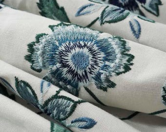May include: Close-up of a white fabric with blue and green embroidered floral designs. The fabric has a textured surface and the flowers are detailed with multiple shades of blue.