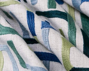 May include: A textured off-white fabric featuring an embroidered pattern of abstract, thick, curved lines in various shades of blue, teal, and light green. The embroidery creates a raised, tactile surface across the woven material.