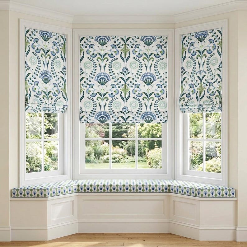 May include: Three Roman shades featuring a white background with a repeating blue and green floral and botanical pattern. A matching upholstered bench cushion with a smaller blue and green geometric print sits below the windows.