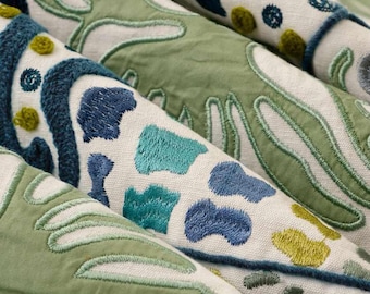 May include: A textile fabric featuring intricate embroidery in shades of sage green, navy blue, and teal. The design consists of organic, flowing shapes and small circular dots stitched onto a cream-colored woven base material.