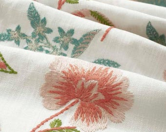 May include: A white fabric featuring an embroidered floral pattern with large, textured peach-colored blossoms and smaller, detailed green leaves and stems. The material has visible woven threads and is arranged in soft, flowing folds across the surface.