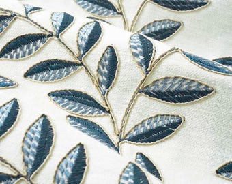 May include: Off-white fabric featuring a repeating pattern of embroidered dark teal leaves. The leaves are arranged in branching stems with a textured stitch detail, outlined in a light beige thread that contrasts against the cream background.