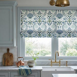 May include: A Roman shade featuring a repeating botanical pattern in shades of blue, green, and white. The fabric displays stylized floral and leaf motifs arranged in a symmetrical vertical layout, covering the upper portion of a window.