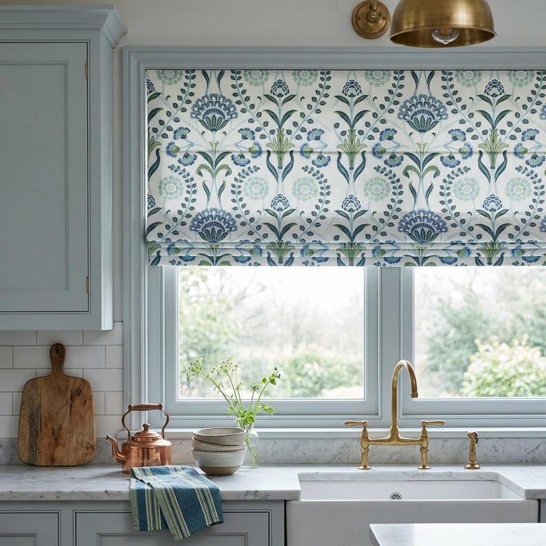 May include: A Roman shade featuring a repeating botanical pattern in shades of blue, green, and white. The fabric displays stylized floral and leaf motifs arranged in a symmetrical vertical layout, covering the upper portion of a window.