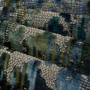 May include: Close-up of a textured fabric with a pattern of overlapping shapes in shades of blue, green, and gold. The fabric has a woven appearance with a mix of smooth and textured areas, creating a layered effect. The overall aesthetic is modern and sophisticated.