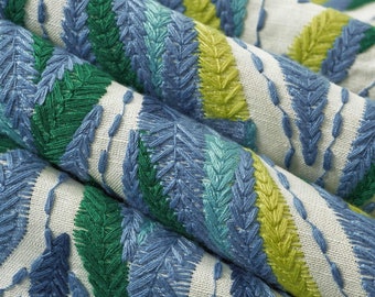 May include: A light-colored fabric featuring dense, textured embroidery in shades of blue, emerald green, and lime green. The stitching forms repeating diagonal leaf-like patterns, creating a raised, tactile surface with visible thread details.