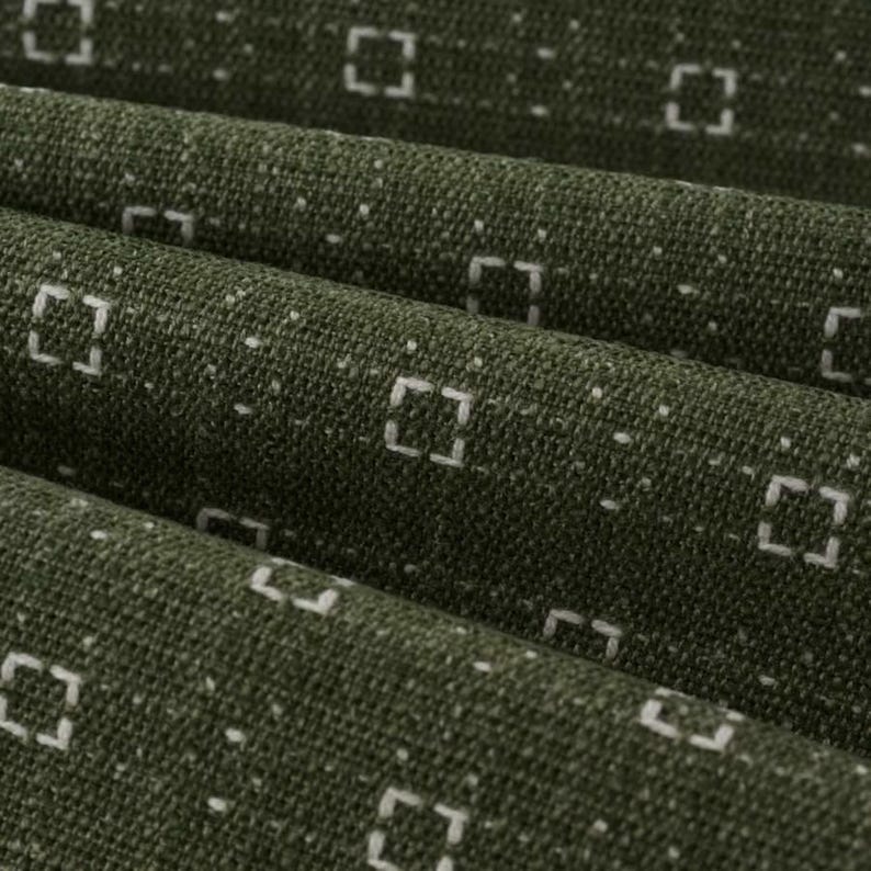 May include: A dark olive green fabric featuring a repeating pattern of small, white, dashed square outlines arranged in horizontal rows. The textile has a visible woven texture with subtle folds creating depth across the surface of the material.
