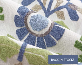 May include: A white fabric featuring dense embroidery in shades of blue, light green, and tan. The design consists of stylized floral and leaf motifs with textured stitching. A blue rectangular overlay displays the text BACK IN STOCK!