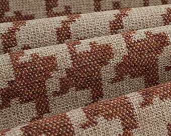 May include: A woven fabric featuring a repeating houndstooth pattern in deep rust orange and light beige. The material has a visible textured weave with a coarse, durable appearance, showing folds that create depth across the surface of the textile.
