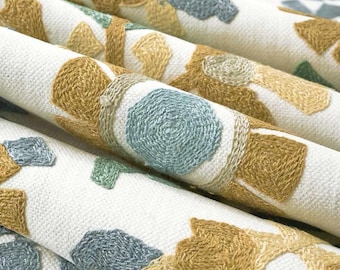 May include: Off-white fabric featuring an embroidered pattern of repeating geometric shapes in mustard yellow and muted blue-green. The textured embroidery creates a raised, tactile surface across the folded textile material.