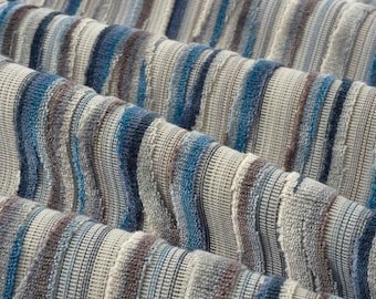 May include: A textile fabric featuring a vertical striped pattern in shades of blue, gray, and white. The material has a textured, ribbed surface with alternating smooth and raised woven sections that create a dimensional, wavy appearance.