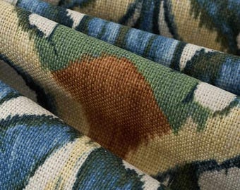 May include: A woven fabric featuring a textured surface with a repeating pattern of blue, green, and tan shapes. The material displays a visible grid-like weave structure with folds that create depth and shadows across the colorful printed design.