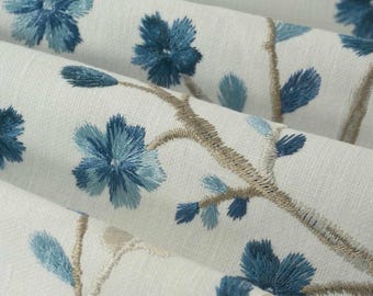 May include: A white fabric featuring an embroidered pattern of blue flowers and light brown stems. The floral design consists of five-petaled blossoms in varying shades of blue, connected by thin, textured branches that create a repeating motif.