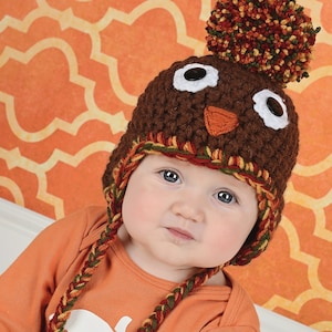 May include: A baby wearing a brown crocheted turkey hat with a brown felt beak and white felt eyes. The hat has a brown, orange, and yellow pom-pom on top.