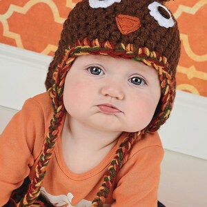 May include: A baby wearing a brown crocheted turkey hat with a brown pom-pom on top. The hat has white eyes, a brown beak, and a red and yellow braided strap. The baby is wearing an orange long-sleeved shirt and blue jeans.