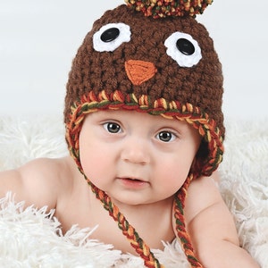 May include: A baby wearing a brown crocheted turkey hat with a colorful pom-pom on top. The hat has white eyes, a brown beak, and a red and orange wattle. The baby is lying on a white fluffy blanket.