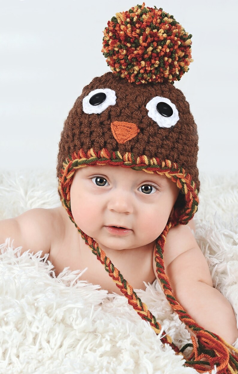 May include: A baby wearing a brown crocheted turkey hat with a colorful pom-pom on top. The hat has white eyes, a brown beak, and a red and orange wattle. The baby is lying on a white fluffy blanket.